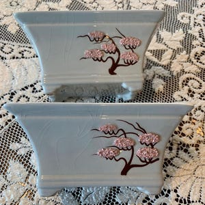May include: Two rectangular ceramic planters with a light blue crackle glaze finish. Each planter features a dark brown painted tree branch design with raised, textured pink floral clusters on the front side and four small feet at the base.