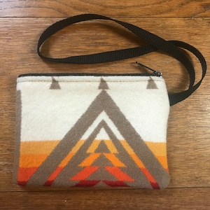 May include: A rectangular, zippered pouch with a black strap. The pouch features a geometric design in shades of white, gray, orange, and brown. The design resembles a mountain range. The pouch is likely made of a soft, textured fabric.