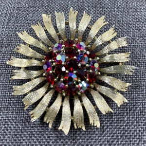 May include: A gold tone brooch with a large red rhinestone cluster in the center. The brooch is shaped like a flower with many petals.