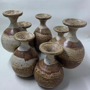 May include: A collection of six ceramic vases with a unique design. The vases have a bulbous base, a slender neck, and a flared top. The color scheme includes shades of brown, white, and speckled accents, creating a rustic aesthetic.