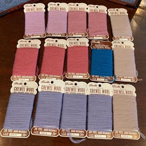 May include: A collection of spools of Bucilla Crewel Wool in various shades of pink, purple, and blue. Each spool is labeled with details including the yardage, color number, and the text "Hand Washable".