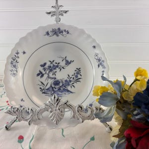 May include: White porcelain plate with a blue floral design, displayed on a decorative white metal stand. The plate has a scalloped edge. A bouquet of colorful flowers is in the background.