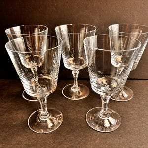May include: Five clear, stemmed wine glasses with etched vertical lines and a small, stemmed glass inside each. The glasses have a classic design with a round base and a slender stem. The background is a dark brown.