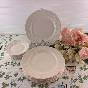 May include: A collection of off-white ceramic plates with a floral embossed design. The plates are stacked and displayed on a floral patterned tablecloth, with a decorative plate holder and a bouquet of pink flowers in the background.