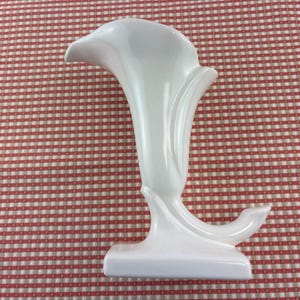 May include: A white ceramic vase in the shape of a calla lily. The vase has a flared opening and a curved stem that extends to a flat base. The vase is smooth and glossy, with a clean, modern design. The vase is approximately 10 inches tall.