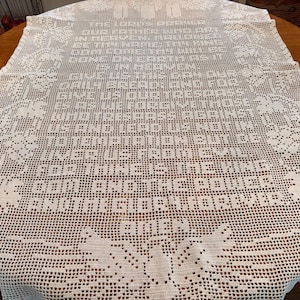 May include: A white, crocheted tablecloth with a detailed pattern and text. The text is a religious passage, and the tablecloth is laid out on a wooden table. The edges of the tablecloth have a decorative floral design.