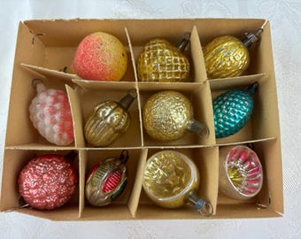 May include: A vintage Christmas ornament set in a cardboard box. The ornaments are various shapes, including spheres, hearts, and teardrops, in colors like gold, red, and teal. Some have textured surfaces, and all have metal caps for hanging.