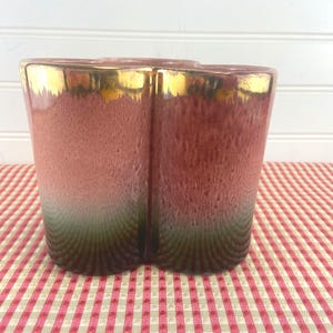 May include: A vintage ceramic vase with a unique shape and a gradient color scheme. The vase transitions from a deep green base to a reddish-pink middle, topped with a gold rim. The vase is sitting on a red and white checkered surface.