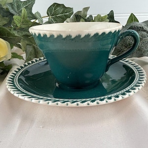 May include: A teal teacup and saucer set with a white scalloped rim. The teacup has a handle and sits on a matching saucer. The set is placed on a white tablecloth with yellow roses and greenery in the background.