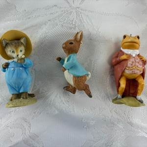 May include: Three anthropomorphic animal figurines: a cat wearing a yellow hat and blue overalls, a brown rabbit in a blue jacket, and a yellow frog dressed in a red coat with a white cravat and a patterned vest, all standing in a row.