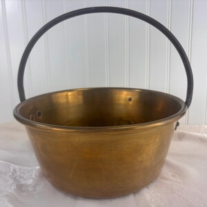 May include: A vintage brass cauldron with a black metal handle. The cauldron has a rounded bowl shape and is made of shiny, golden-colored metal. It is likely used for cooking or decorative purposes.