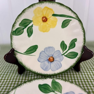 May include: A white ceramic plate with a scalloped edge, hand-painted with a yellow and a blue flower, green leaves, and a green border. The plate is displayed on a dark brown stand against a green and white checkered background.
