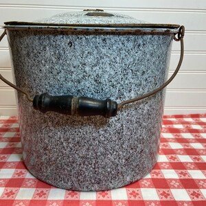 May include: Vintage speckled enamelware bucket with lid. The bucket has a gray and white speckled pattern, a metal handle with a wooden grip, and a matching lid. The bucket is sitting on a red and white checkered tablecloth.