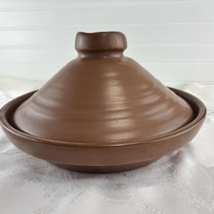 May include: A brown ceramic tagine cooking vessel consisting of a wide, shallow circular base and a tall, conical lid with a knob handle at the top. The exterior surface of the cookware features a smooth, matte finish with subtle horizontal ridges.