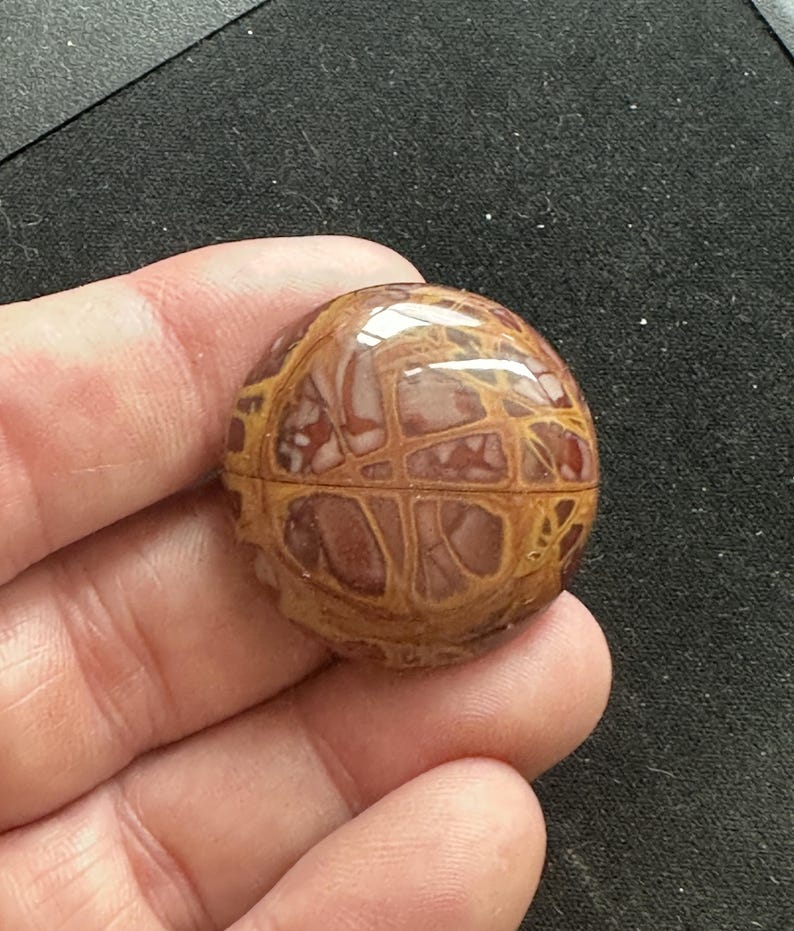 May include: A round, polished cabochon of brown and tan jasper with a swirling, abstract pattern.