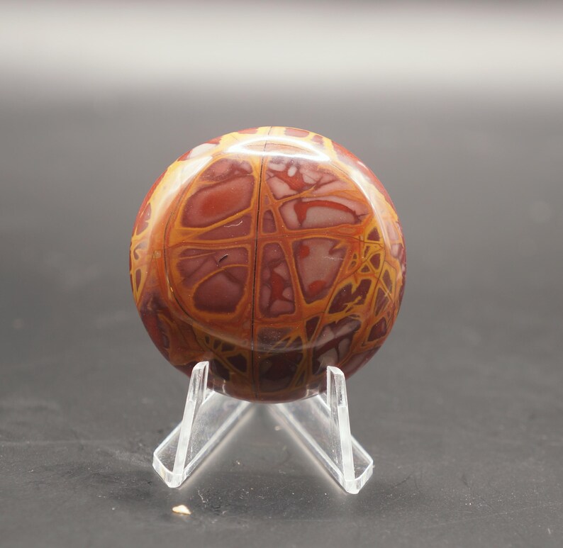 May include: A round, polished stone with a red, orange, and white swirling pattern. The stone is displayed on a clear plastic stand.