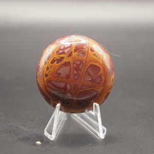 May include: A polished red and yellow agate stone with a unique pattern, displayed on a clear acrylic stand.