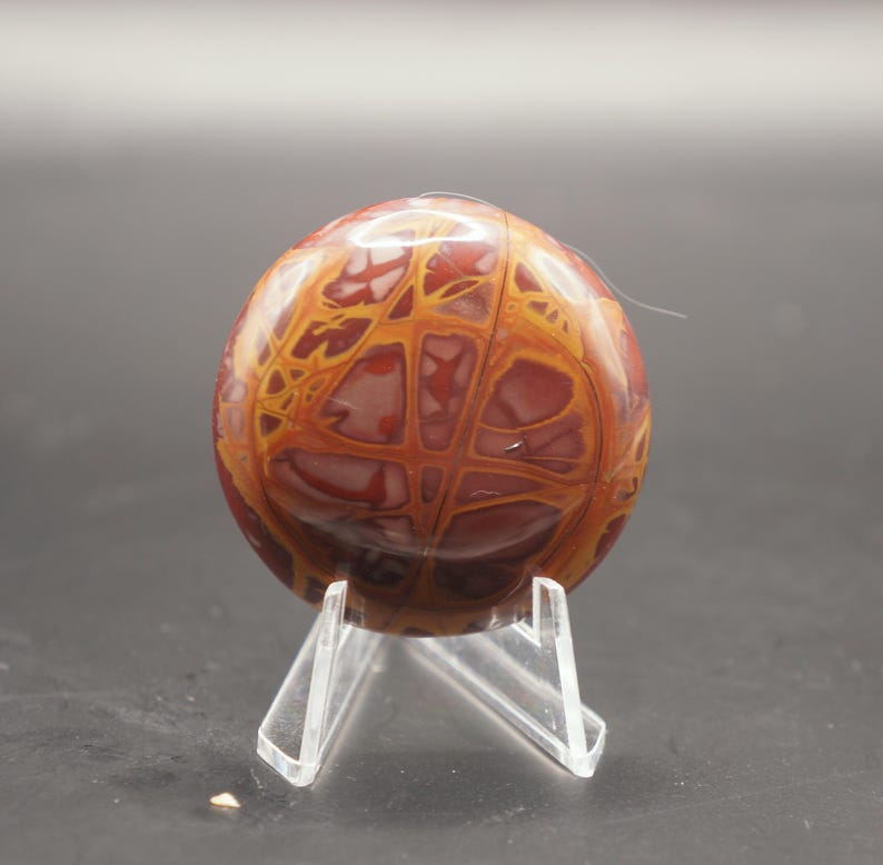 May include: A polished red and yellow agate stone with a unique pattern, displayed on a clear acrylic stand.