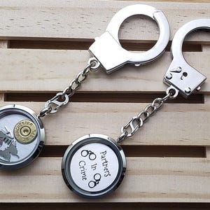 May include: Two silver-toned metal keychains featuring miniature handcuffs attached to circular lockets. One locket contains a Winchester 38 SPL bullet casing and a pink crystal, while the other displays the text Partners in Crime with handcuff icons.