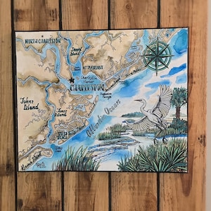 May include: Watercolor map of Charleston, South Carolina, featuring a compass rose and a flying bird. The map includes place names like North Charleston, Johns Island, and the Atlantic Ocean. The artwork is framed and mounted on a wooden background.