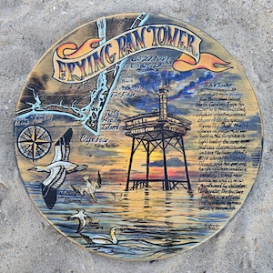 May include: A round wooden sign with a hand-painted coastal scene. The sign features a map, a compass rose, and the text "FRYING PAN TOWER." It depicts a tower over the ocean with birds flying above the water. The sign is on a sandy surface.