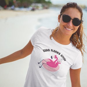 Puede incluir: Camiseta blanca con un flamenco rosa en un flotador de piscina sosteniendo una bebida. El texto "Zero Flocks Given" está impreso en la camiseta.