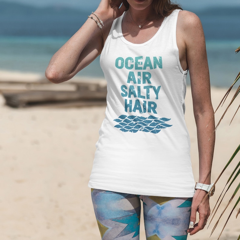 May include: White tank top with the text "Ocean Air Salty Hair" in blue and green lettering. The text is stylized with a wavy line underneath. The tank top is being worn by a person on a beach.