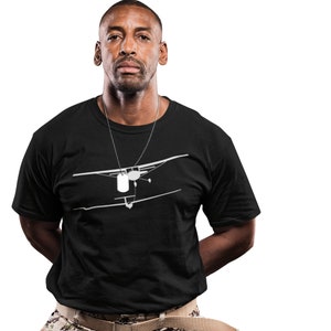 May include: A black t-shirt with a white silhouette of a small airplane with two dog tags hanging from it. The person wearing the shirt is a man with short black hair and a beard.