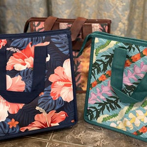 May include: Three tote bags with floral patterns. The front bag has a navy blue background with large pink and white hibiscus flowers and dark blue handles. The middle bag has a brown background with pink flowers. The third bag has a teal background with floral garlands.