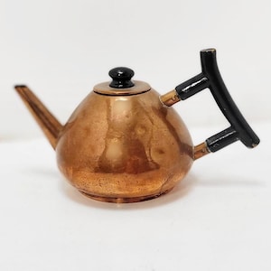 May include: A small copper teapot with a black handle and knob. The teapot has a rounded body and a spout.