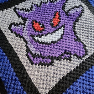 Puede incluir: Manta de ganchillo con una imagen pixelada en morado y blanco de un fantasma con ojos rojos. La manta tiene un borde azul, negro y gris.