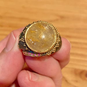 May include: A statement ring featuring a large, round, translucent gemstone with golden inclusions, set in a dark metal band with gold accents. Small pink gemstones are set along the bottom of the ring.