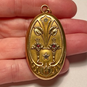 May include: A gold oval locket with a floral design and red and white gemstones. The locket has a loop at the top for a chain.