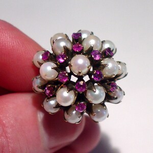 May include: A cluster ring featuring a floral design. The ring is adorned with numerous white pearls and vibrant pink gemstones, set in a gold-toned metal. The ring's design is intricate and eye-catching, showcasing a classic jewelry style.
