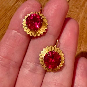 May include: A pair of gold-toned earrings featuring round, faceted, vibrant red stones set in the center. Each stone is surrounded by a textured, sunburst-style metal frame with radiating lines, and each earring includes a small metal hook attachment.