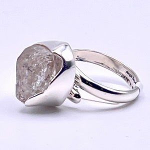 May include: A silver ring with a large, rough-cut, clear crystal. The crystal is set in a polished silver bezel. The ring band is also silver and has a simple design. The ring is shown against a white backdrop. The ring is a piece of jewellery.