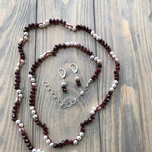 Op de afbeelding: Een ketting en bijpassende oorbellen set met afwisselend witte parels en dieprode kralen, geregen op een rood koord. De ketting heeft een zilveren sluiting en verlengketting. De oorbellen hebben zilveren haken. De sieraden worden getoond op een houten oppervlak.