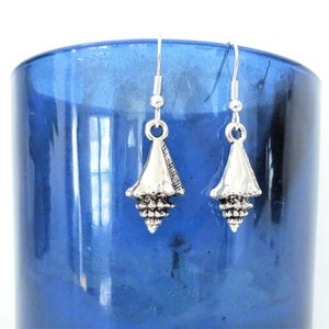 May include: A pair of silver-colored seashell earrings. Each earring features a detailed, cone-shaped shell charm suspended from a simple hook. The earrings are displayed against a blurred blue background, highlighting their intricate design and nautical theme.