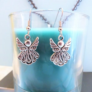 Puede incluir: Un par de pendientes de &aacute;ngel de plata con un dise&ntilde;o detallado. Los pendientes cuelgan de un frasco de vidrio lleno de l&iacute;quido azul verdoso.