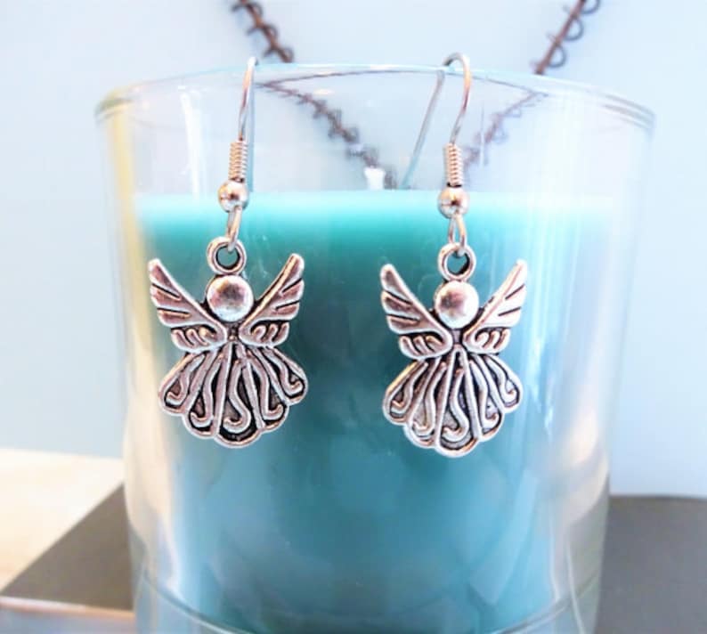 Puede incluir: Un par de pendientes de &aacute;ngel de plata con un dise&ntilde;o detallado. Los pendientes cuelgan de un frasco de vidrio lleno de l&iacute;quido azul verdoso.