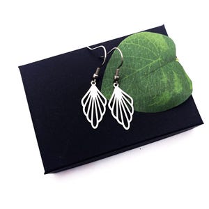 May include: A pair of silver-colored, openwork earrings in a fan or leaf design. The earrings are displayed on a black box with a green leaf, set against a white background. The earrings have a hook closure.