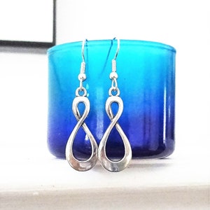 May include: A pair of silver infinity symbol earrings with a drop design. The earrings are hanging from a blue and purple ombre background.