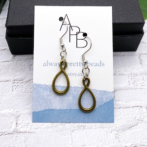 May include: A pair of bronze-colored infinity symbol earrings with silver-colored ear wires. The earrings are displayed on a white card with the text "always pretty beads" and a blue watercolor design. The background includes a black box and a white brick wall.