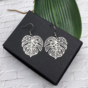 May include: A pair of silver earrings shaped like monstera leaves. The earrings are delicate and intricate, with a cutout design that shows the veins of the leaves. The earrings are hanging from a black box.