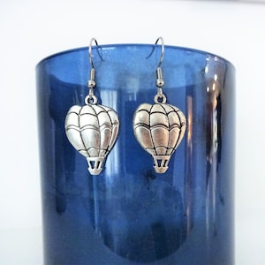 May include: Silver hot air balloon dangle earrings with a silver hook. The earrings are hanging from a blue glass container.
