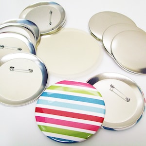 May include: A set of blank round button badges with a colorful striped button badge. The blank badges are silver with a pin back. The striped button badge has a blue, pink, red, and green striped design.