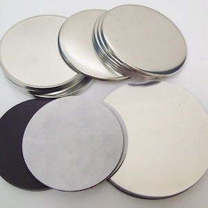 May include: A collection of shiny, silver-colored, round metal discs. Some discs are stacked, while others are separate. The image also shows black and white circular materials, possibly for crafts or DIY projects.