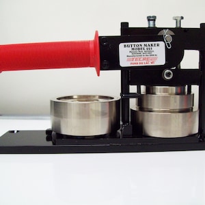 May include: A black button maker with a red handle. The machine has a label that reads "Button Maker Model 925 Patent No. 6,831,927 Manufactured in the USA by Tecre Fond du Lac, WI".