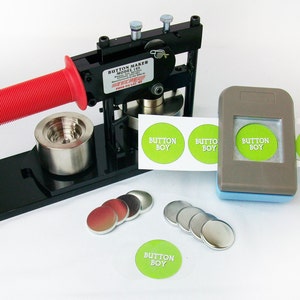 Op de afbeelding: Een zwarte knopenmaker met een rode handgreep, een rol groene en witte "Button Boy" stickers, een grijze knopenpons en een stapel zilveren en rode knopenruggen.