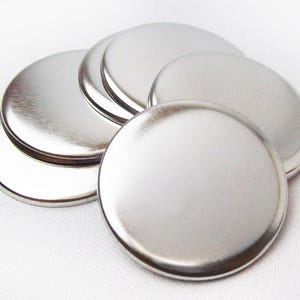 May include: A collection of shiny, silver-colored, circular metal blanks. The blanks are arranged in a slightly overlapping pattern, with a few stacked together. The surfaces reflect light, creating highlights and shadows. These are suitable for crafting.